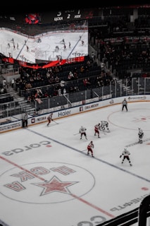 A professional ice hockey game is in progress inside an arena, with players in red and white jerseys actively engaging on the rink. The scoreboard above displays the score and game details, while spectators are seated in the stands. A large screen shows a live feed of the match action.