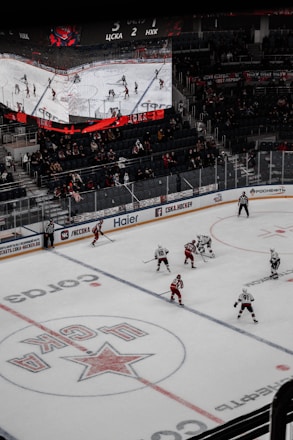 A professional ice hockey game is in progress inside an arena, with players in red and white jerseys actively engaging on the rink. The scoreboard above displays the score and game details, while spectators are seated in the stands. A large screen shows a live feed of the match action.