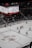 A professional ice hockey game is in progress inside an arena, with players in red and white jerseys actively engaging on the rink. The scoreboard above displays the score and game details, while spectators are seated in the stands. A large screen shows a live feed of the match action.