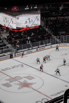 A professional ice hockey game is in progress inside an arena, with players in red and white jerseys actively engaging on the rink. The scoreboard above displays the score and game details, while spectators are seated in the stands. A large screen shows a live feed of the match action.