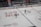 Two ice hockey players wearing helmets and uniforms are on an ice rink. The rink has various advertising logos, and there's a boundary wall separating the rink from a seating area with spectators. Some staff members are visible behind the wall.