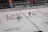 Two ice hockey players wearing helmets and uniforms are on an ice rink. The rink has various advertising logos, and there's a boundary wall separating the rink from a seating area with spectators. Some staff members are visible behind the wall.