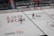Two ice hockey players wearing helmets and uniforms are on an ice rink. The rink has various advertising logos, and there's a boundary wall separating the rink from a seating area with spectators. Some staff members are visible behind the wall.