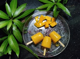 Three yellow mango popsicles and sliced mango pieces rest on a clear, textured plate surrounded by vibrant green mango leaves. The background is a dark surface, enhancing the vividness of the colors.