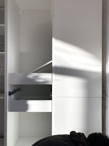 A white cabinet with multiple compartments, including a partially open drawer. Soft natural light creates shadows and highlights on the surface. A person is partially visible at the bottom right corner, possibly assembling or inspecting the cabinet.
