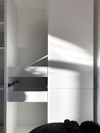 A white cabinet with multiple compartments, including a partially open drawer. Soft natural light creates shadows and highlights on the surface. A person is partially visible at the bottom right corner, possibly assembling or inspecting the cabinet.