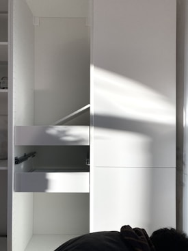 A white cabinet with multiple compartments, including a partially open drawer. Soft natural light creates shadows and highlights on the surface. A person is partially visible at the bottom right corner, possibly assembling or inspecting the cabinet.