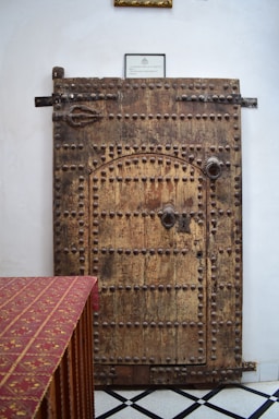 A large, rustic wooden door with metal studs is set against a neutral wall. A piece of furniture with a red patterned cloth draped over it is positioned on the left. The floor is tiled with a black and white diamond pattern, and a small framed sign hangs above the door.