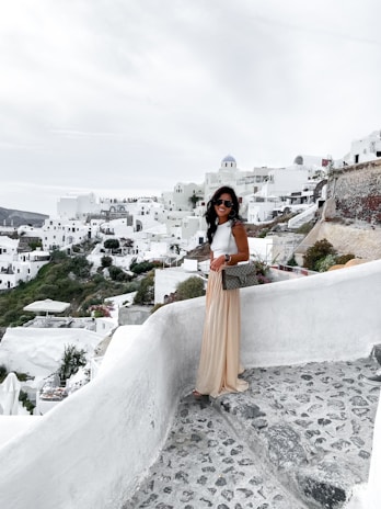 A woman stands on a stone pathway overlooking a scenic view of white buildings on a hillside, typical of Cycladic architecture. The sky is overcast, creating a calm and subdued atmosphere. She is dressed in a white top, long beige skirt, and sunglasses, carrying a bag.