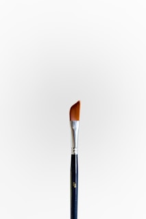 A single paintbrush with a silver ferrule and angled bristles stands upright against a plain white background. The handle is dark with a metallic finish at the top.