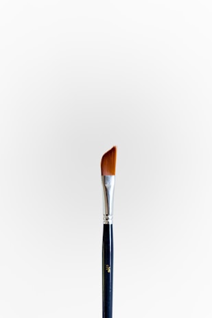 A single paintbrush with a silver ferrule and angled bristles stands upright against a plain white background. The handle is dark with a metallic finish at the top.