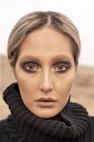 A close-up portrait of a person with intense eye makeup, featuring dark smoky eyes and shimmering highlights. The makeup is complemented by a neutral, matte complexion and muted lipstick. The person is wearing a textured black knitted garment, with the background blurred, giving an impression of an outdoor setting.
