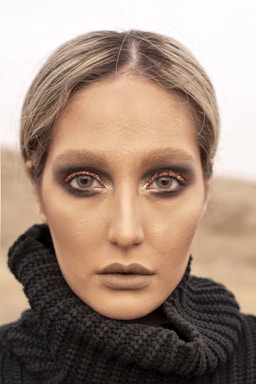 A close-up portrait of a person with intense eye makeup, featuring dark smoky eyes and shimmering highlights. The makeup is complemented by a neutral, matte complexion and muted lipstick. The person is wearing a textured black knitted garment, with the background blurred, giving an impression of an outdoor setting.