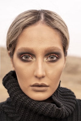 A close-up portrait of a person with intense eye makeup, featuring dark smoky eyes and shimmering highlights. The makeup is complemented by a neutral, matte complexion and muted lipstick. The person is wearing a textured black knitted garment, with the background blurred, giving an impression of an outdoor setting.
