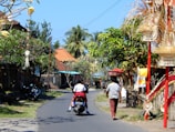 A narrow, winding street lined with colorful traditional Indonesian decorations and lush green trees. A person rides a motorcycle with a passenger down the sunlit road, while another person walks nearby carrying a basket on their head. Traditional Balinese offerings and structures adorn the roadside, adding vibrant cultural elements to the scene. The sky is clear blue, and tropical plants are visible.