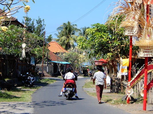 A narrow, winding street lined with colorful traditional Indonesian decorations and lush green trees. A person rides a motorcycle with a passenger down the sunlit road, while another person walks nearby carrying a basket on their head. Traditional Balinese offerings and structures adorn the roadside, adding vibrant cultural elements to the scene. The sky is clear blue, and tropical plants are visible.