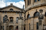 Stone busts of historical figures are mounted on tall pedestals in front of an ornate building with classical architecture. The building is adorned with large windows and decorative stonework. A fence runs along the front of the structure.