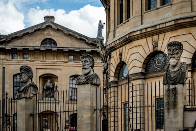 Stone busts of historical figures are mounted on tall pedestals in front of an ornate building with classical architecture. The building is adorned with large windows and decorative stonework. A fence runs along the front of the structure.
