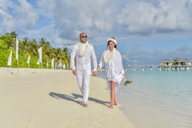 A couple dressed in white, adorned with floral garlands, is walking hand in hand along a tropical beach. The beach is lined with palm trees and white flags, and a wooden pier extends into the turquoise sea. The couple appears joyful, with the sun casting soft shadows on the sand.