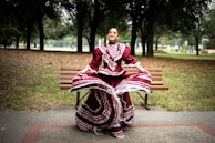 A woman is gracefully dancing in a park while wearing a traditional maroon and white folkloric dress with intricate patterns. She is standing in front of a wooden bench surrounded by trees under a cloudy sky, which creates a serene and festive atmosphere.