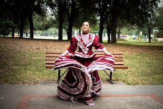 A woman is gracefully dancing in a park while wearing a traditional maroon and white folkloric dress with intricate patterns. She is standing in front of a wooden bench surrounded by trees under a cloudy sky, which creates a serene and festive atmosphere.