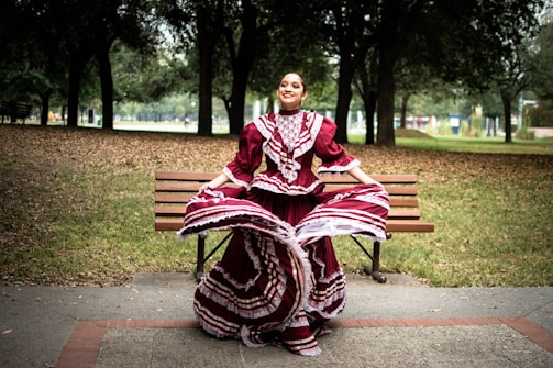 A woman is gracefully dancing in a park while wearing a traditional maroon and white folkloric dress with intricate patterns. She is standing in front of a wooden bench surrounded by trees under a cloudy sky, which creates a serene and festive atmosphere.