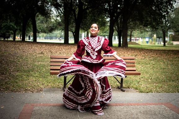 A woman is gracefully dancing in a park while wearing a traditional maroon and white folkloric dress with intricate patterns. She is standing in front of a wooden bench surrounded by trees under a cloudy sky, which creates a serene and festive atmosphere.