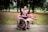 A woman is gracefully dancing in a park while wearing a traditional maroon and white folkloric dress with intricate patterns. She is standing in front of a wooden bench surrounded by trees under a cloudy sky, which creates a serene and festive atmosphere.