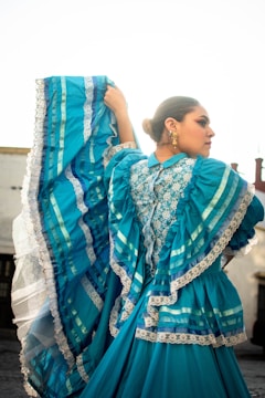 A person is dressed in a traditional vibrant blue dress adorned with intricate lace and patterns. The dress features multiple layers and ruffles, creating a flowing effect. The person is holding up part of the dress, showcasing its texture and details. The hairstyle is a neat bun, and they are wearing gold earrings, adding to the elegance of the attire. The background is bright and slightly blurred, enhancing the focus on the dress.