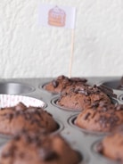 Freshly baked chocolate muffins in a muffin tin, topped with chunks of chocolate. Some of the muffin slots are empty, and there is a small decorative flag featuring a cartoon cake.
