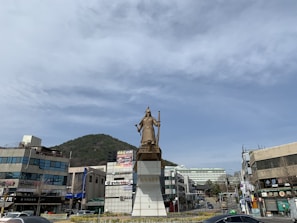 A large statue of a historical figure stands prominently on a decorated pedestal in an urban setting. The backdrop features modern buildings, hillside greenery, and a wide, partly cloudy sky.