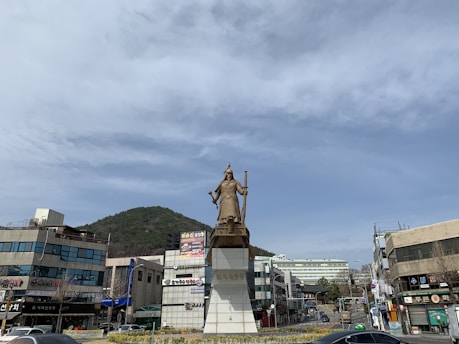A large statue of a historical figure stands prominently on a decorated pedestal in an urban setting. The backdrop features modern buildings, hillside greenery, and a wide, partly cloudy sky.
