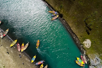 Aerial view of a vibrant turquoise river with multiple colorful boats lined along the shoreline. The boats are painted in bright hues such as yellow, red, and blue. The water appears clear, and the surrounding area includes a grassy, rocky bank with visible shadows.