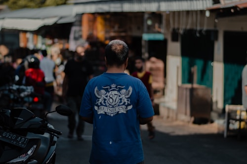 A man wearing a blue t-shirt with the number 87 and the word Outsiders printed on the back stands in what appears to be a bustling street market. Surrounding him are various blurred figures and a motorcycle. The background comprises market stalls and some buildings.