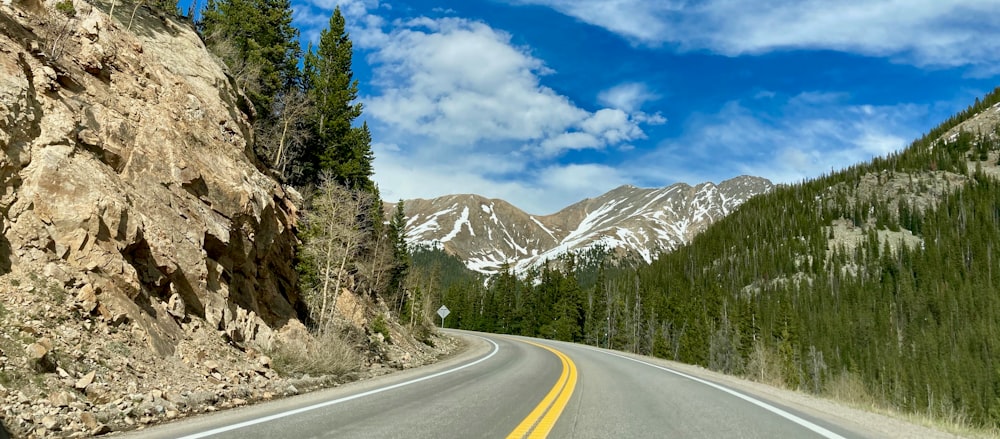 gray concrete road between green trees and white mountains under blue sky and white clouds during