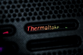 A close-up view of a textured black surface with the brand name 'Thermaltake' illuminated in a gradient of colors ranging from red to yellow. The design features evenly spaced horizontal slots with glowing accents.