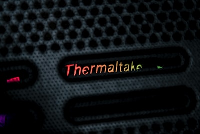 A close-up view of a textured black surface with the brand name 'Thermaltake' illuminated in a gradient of colors ranging from red to yellow. The design features evenly spaced horizontal slots with glowing accents.