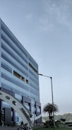 A modern, multi-story office building with a geometric design and large windows. The structure incorporates blue and metallic tones, with a distinctive arch near its base. Nearby, palm trees and manicured plants are visible, adding a touch of greenery. A clear sky suggests a pleasant day.