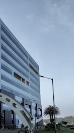 A modern, multi-story office building with a geometric design and large windows. The structure incorporates blue and metallic tones, with a distinctive arch near its base. Nearby, palm trees and manicured plants are visible, adding a touch of greenery. A clear sky suggests a pleasant day.