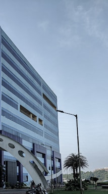 A modern, multi-story office building with a geometric design and large windows. The structure incorporates blue and metallic tones, with a distinctive arch near its base. Nearby, palm trees and manicured plants are visible, adding a touch of greenery. A clear sky suggests a pleasant day.