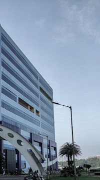 A modern, multi-story office building with a geometric design and large windows. The structure incorporates blue and metallic tones, with a distinctive arch near its base. Nearby, palm trees and manicured plants are visible, adding a touch of greenery. A clear sky suggests a pleasant day.