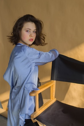 A person with medium-length dark hair is standing next to a director's chair, wearing a silky blue robe or garment. The background appears to be a plain tan or beige wall with natural lighting creating shadows.