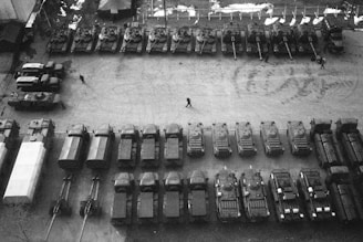An aerial view of a lot filled with military vehicles, including tanks and trucks, arranged in neat rows. There are a few people walking between the vehicles, and some tire tracks are visible on the ground. The scene is monochromatic, emphasizing the military equipment and the structured organization of the layout.