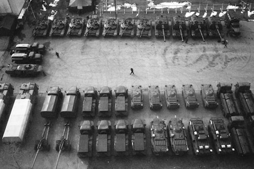 An aerial view of a lot filled with military vehicles, including tanks and trucks, arranged in neat rows. There are a few people walking between the vehicles, and some tire tracks are visible on the ground. The scene is monochromatic, emphasizing the military equipment and the structured organization of the layout.