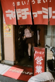 A person is entering a small shop or restaurant with red fabric signs displaying white Japanese text. A red welcome mat is placed at the entrance with more Japanese writing. The person is carrying a light-colored backpack with a small plush toy attached. A scooter is parked nearby.