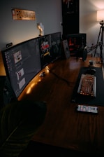 A modern gaming setup featuring a large curved monitor displaying a game interface. The desk includes a mechanical keyboard with RGB lighting, a microphone, and a cup of coffee. There is a smartphone on the table, and speakers on either side of the monitor. A soft lamp light provides ambient lighting, and a decorative sign is mounted on the wall.