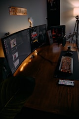 A modern gaming setup featuring a large curved monitor displaying a game interface. The desk includes a mechanical keyboard with RGB lighting, a microphone, and a cup of coffee. There is a smartphone on the table, and speakers on either side of the monitor. A soft lamp light provides ambient lighting, and a decorative sign is mounted on the wall.