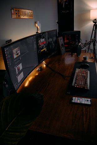 A modern gaming setup featuring a large curved monitor displaying a game interface. The desk includes a mechanical keyboard with RGB lighting, a microphone, and a cup of coffee. There is a smartphone on the table, and speakers on either side of the monitor. A soft lamp light provides ambient lighting, and a decorative sign is mounted on the wall.