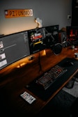 A computer desk setup featuring dual monitors, a keyboard with red backlighting, a microphone, a gaming controller, and a cup of coffee. One monitor displays a game, Call of Duty: Cold War, while the other screen shows a chat application. The desk is made of wood, and there is an orange 'UNIMOG' sign on the wall above the monitors. The desk also has a smartphone, a small speaker, and other small desk items.
