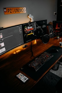 A computer desk setup featuring dual monitors, a keyboard with red backlighting, a microphone, a gaming controller, and a cup of coffee. One monitor displays a game, Call of Duty: Cold War, while the other screen shows a chat application. The desk is made of wood, and there is an orange 'UNIMOG' sign on the wall above the monitors. The desk also has a smartphone, a small speaker, and other small desk items.
